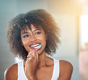 Woman with brown curly hair smiling while brushing her teeth in front of the mirror